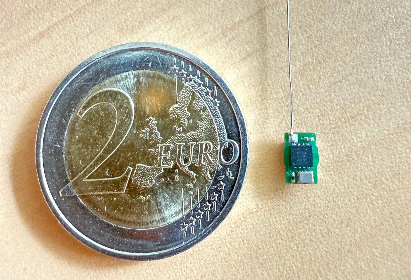Der VeluFinder-Sender liegt neben einem 2-Euro-Stück; die Platine ist
              deutlich kleiner als die Münze, aus ihr ragt ein feiner Antennendraht.